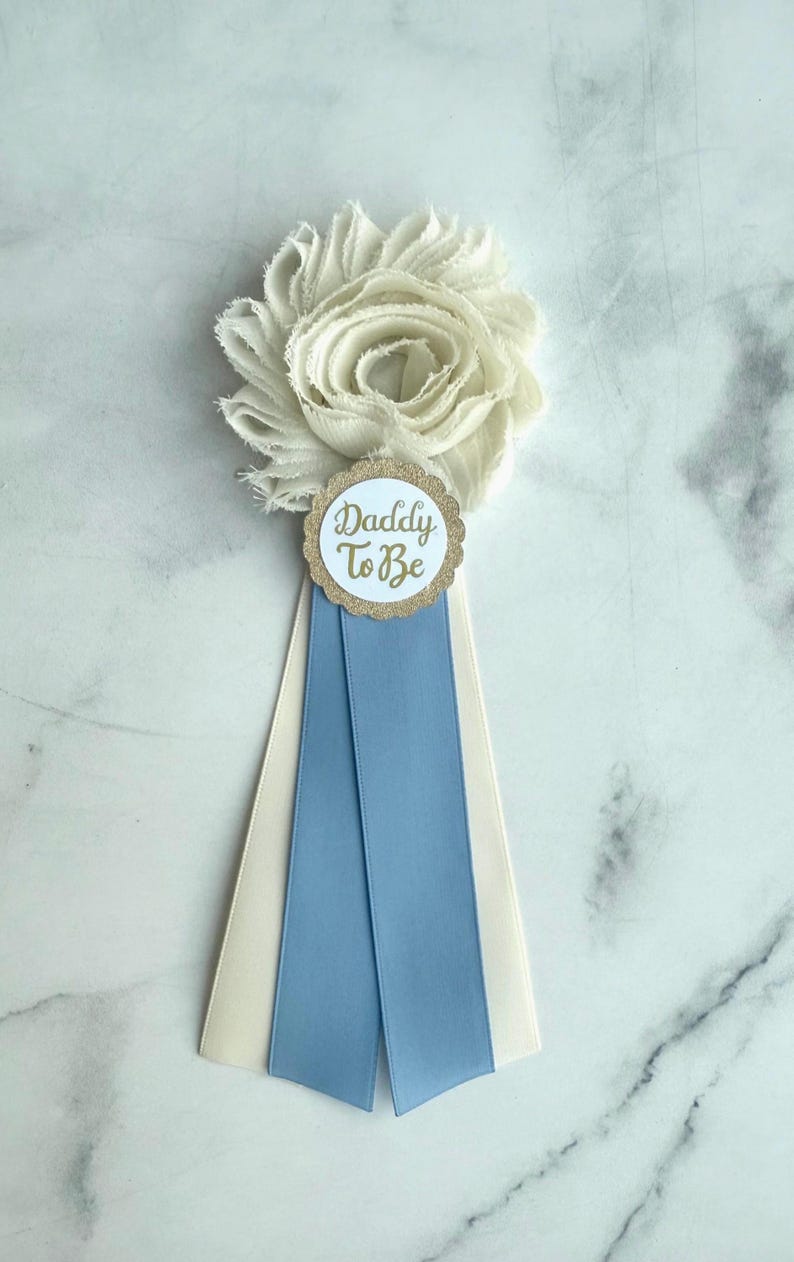 K&ouml;nnte beinhalten: Eine wei&szlig;e Stoffblume mit einem goldumrandeten runden Anh&auml;nger mit der Aufschrift "Daddy To Be". Der Anh&auml;nger ist an einem Band mit einem hellblauen Streifen und einem cremefarbenen Streifen befestigt, f&uuml;r eine Babyparty oder Gender Reveal.