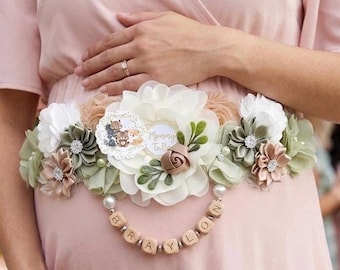 Peut inclure: Une ceinture de maternité florale avec une fleur crème et une chaîne de perles en bois épelant "BRAYLON". La ceinture présente des fleurs en tissu blanches, vertes et beiges, avec un petit emblème rond qui dit "Mommy To Be".
