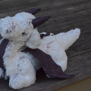 May include: A white plush dragon with a gray fabric wing and a gray fabric horn. The dragon is lying on a wooden surface.