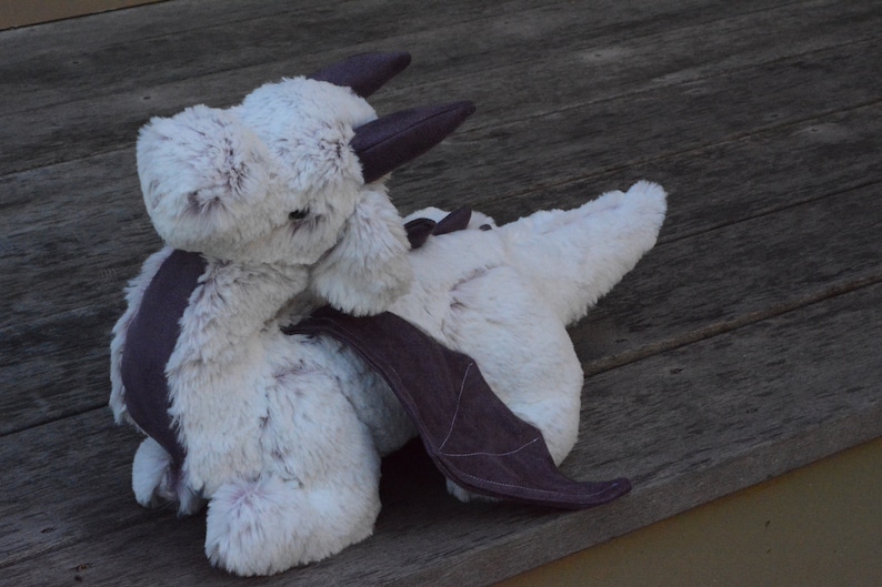 May include: A white plush dragon with a gray fabric wing and a gray fabric horn. The dragon is lying on a wooden surface.