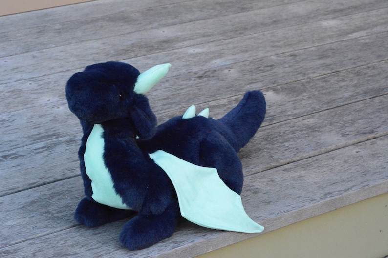 May include: A plush blue dragon with light green wings and horns. The dragon is sitting on a wooden surface.