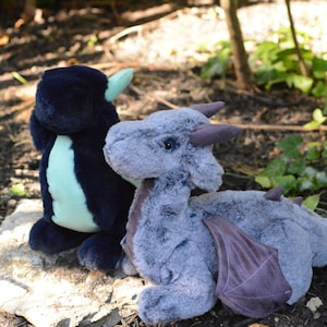May include: Two plush dragon toys, one is dark blue and teal, the other is gray with purple wings. They are sitting on a rock in a garden setting.