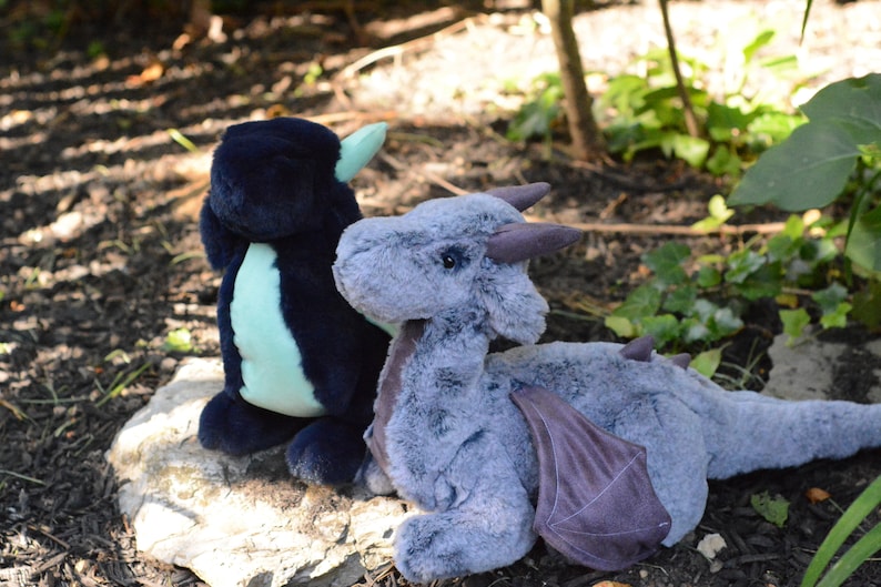 May include: Two plush dragon toys, one is dark blue and teal, the other is gray with purple wings. They are sitting on a rock in a garden setting.