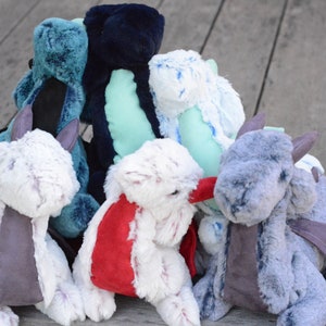 May include: A group of seven plush dragon toys in various colors, including blue, green, white, and gray. The dragons have wings and are made of soft, fuzzy fabric.
