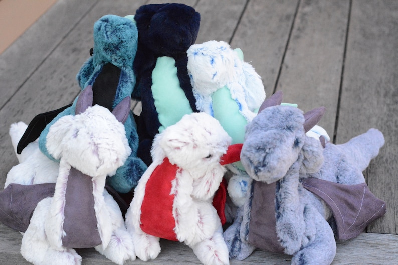 May include: A group of seven plush dragon toys in various colors, including blue, green, white, and gray. The dragons have wings and are made of soft, fuzzy fabric.
