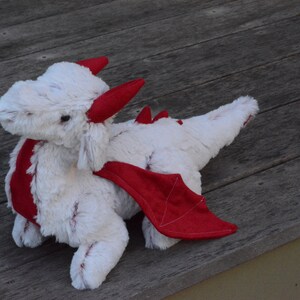 May include: White plush dragon toy with red wings and horns. The dragon is lying on a wooden surface.