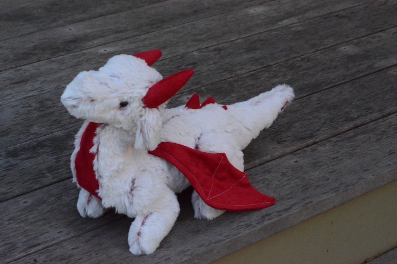 May include: White plush dragon toy with red wings and horns. The dragon is lying on a wooden surface.