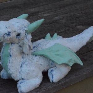 May include: A plush blue and green dragon toy with wings. The dragon has a fluffy white body and a light blue head with a green snout. The wings are a light green color.