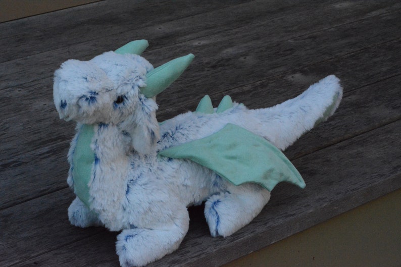 May include: A plush blue and green dragon toy with wings. The dragon has a fluffy white body and a light blue head with a green snout. The wings are a light green color.