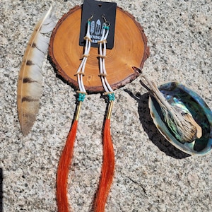 May include: Long, handmade earrings with white cords, turquoise beads, and orange tassels. The earrings are displayed on a wooden slice with a feather and a shell. The earrings have gold-colored metal accents.