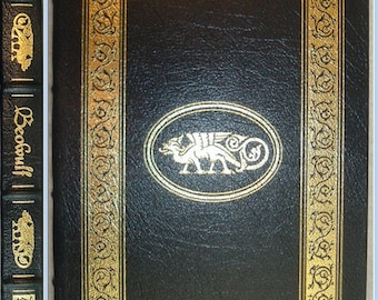 Puede incluir: Un libro de tapa dura negro con detalles dorados. La portada presenta un marco dorado con diseños ornamentados y un óvalo central con una ilustración de dragón. El lomo del libro muestra el título "Beowulf" en dorado.