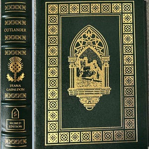 Puede incluir: Libro de tapa dura verde oscuro, "Outlander" de Diana Gabaldón, con detalles dorados. La portada presenta un diseño enmarcado en oro y una ilustración central. El lomo muestra el título, autor y un cardo. Es una edición firmada.