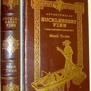 Puede incluir: Un libro de tapa dura marrón titulado Adventures of Huckleberry Finn de Mark Twain. La portada presenta texto dorado en relieve y un gráfico de una persona en un bote. El lomo muestra el título y el autor en letras doradas con adornos.