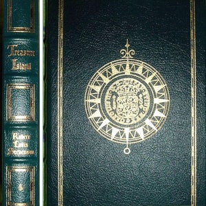Può includere: Un libro con copertina rigida verde scuro intitolato Treasure Island di Robert Louis Stevenson. La copertina presenta un design a bussola e un bordo in rilievo dorato. Il dorso mostra il titolo e l'autore in lettere dorate con cornici.