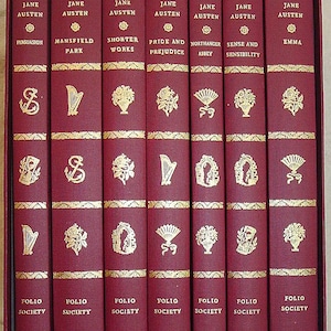 May include: A boxed set of seven burgundy hardcover books by Jane Austen, published by The Folio Society. Each spine features gold-stamped lettering, decorative floral bands, and unique gold icons including harps, fans, and urns.