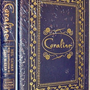 K&ouml;nnte beinhalten: Ein dunkelblaues Hardcover-Buch mit dem Titel "Coraline" von Neil Gaiman, mit goldgepr&auml;gten Schl&uuml;sseln und dem Titel "Coraline". Der Buchr&uuml;cken zeigt den Titel, den Autor und "Signed Edition". Das Buch ist in Zellophan verpackt.
