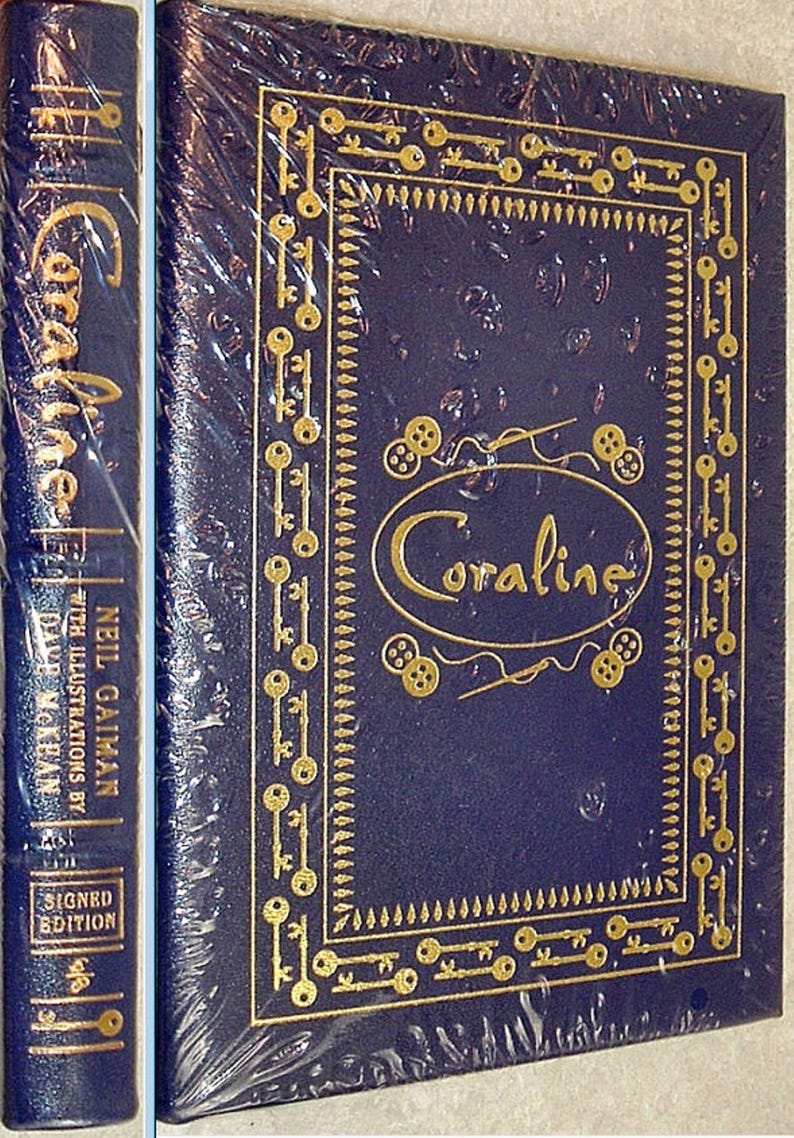 K&ouml;nnte beinhalten: Ein dunkelblaues Hardcover-Buch mit dem Titel "Coraline" von Neil Gaiman, mit goldgepr&auml;gten Schl&uuml;sseln und dem Titel "Coraline". Der Buchr&uuml;cken zeigt den Titel, den Autor und "Signed Edition". Das Buch ist in Zellophan verpackt.