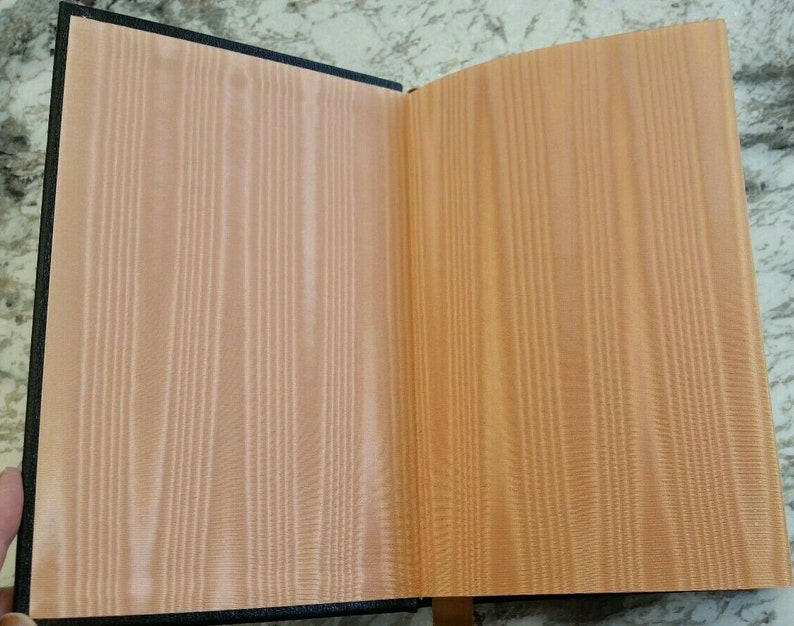 May include: Open book with a black cover and peach-colored pages. The pages have a vertical wood grain pattern. The book is open, revealing the inside pages. The book is on a light-colored surface.