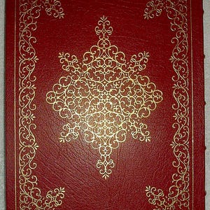May include: A red leather-bound book with gold filigree detailing. The cover features a large, ornate gold design in the center, surrounded by a gold border and corner accents. The book's edges are also gold-trimmed, creating a classic, elegant look.