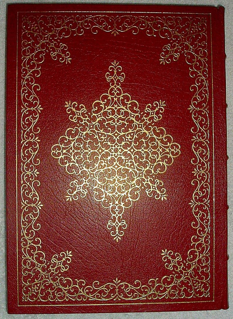 May include: A red leather-bound book with gold filigree detailing. The cover features a large, ornate gold design in the center, surrounded by a gold border and corner accents. The book's edges are also gold-trimmed, creating a classic, elegant look.