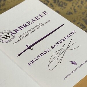 May include: A white book with the title "Warbreaker" in purple lettering. The book is a tenth anniversary edition of the novel, and it is a Dragonsteel Leather Edition. The book is signed by Brandon Sanderson.