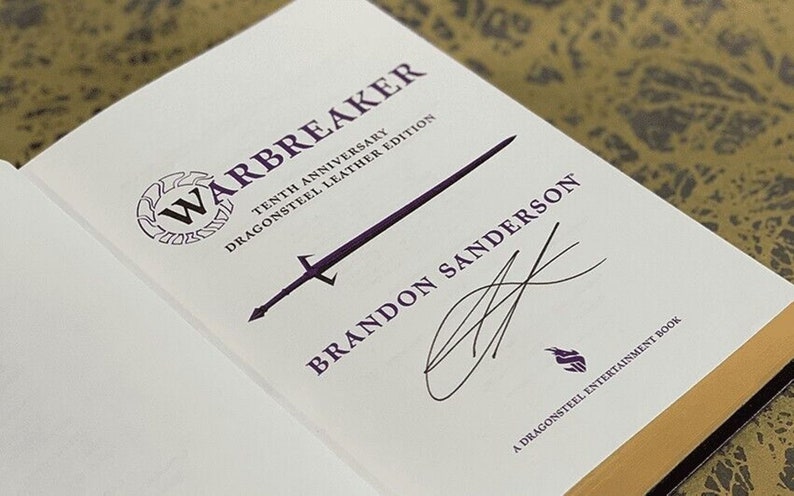 May include: A white book with the title "Warbreaker" in purple lettering. The book is a tenth anniversary edition of the novel, and it is a Dragonsteel Leather Edition. The book is signed by Brandon Sanderson.