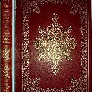 May include: A hardcover book with a deep red cover and gold embossed detailing. The cover features an ornate gold design and a decorative border. The spine displays the title "The Essays of Ralph Waldo Emerson" in gold lettering. The book's overall aesthetic is classic and elegant.