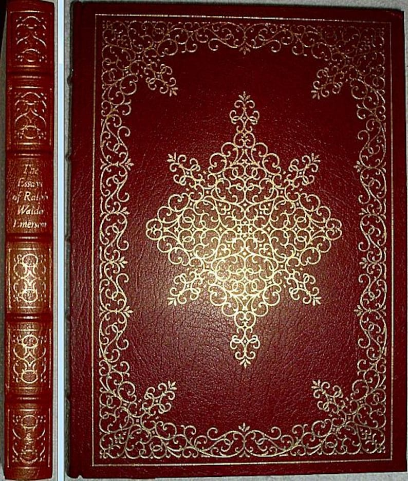 May include: A hardcover book with a deep red cover and gold embossed detailing. The cover features an ornate gold design and a decorative border. The spine displays the title "The Essays of Ralph Waldo Emerson" in gold lettering. The book's overall aesthetic is classic and elegant.