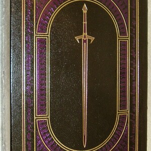 May include: A dark brown leather-bound book with a gold and purple design. The design features a sword in the center and intricate patterns around it.