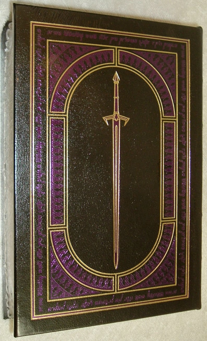 May include: A dark brown leather-bound book with a gold and purple design. The design features a sword in the center and intricate patterns around it.