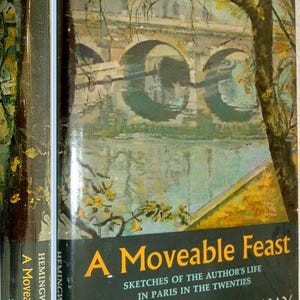 May include: A hardback book titled A Moveable Feast by Ernest Hemingway. The dust jacket features an impressionistic painting of a stone bridge over a river with trees in autumn colours. The title and author name are printed in yellow and white.