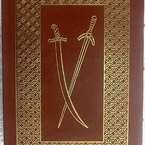 May include: A brown leather-bound book with a gold-colored design on the cover. The design features two crossed swords.