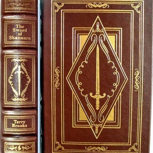Puede incluir: Un libro de tapa dura marrón titulado "The Sword of Shannara" de Terry Brooks. La portada presenta letras doradas en relieve y un diseño en forma de diamante con una espada. El lomo también tiene letras doradas.