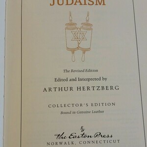 May include: A book titled "JUDAISM" with gold lettering and an illustration of Torah scrolls. The text includes "The Revised Edition," "Edited and Interpreted by ARTHUR HERTZBERG," and "COLLECTOR'S EDITION." The Easton Press, Norwalk, Connecticut is also printed.