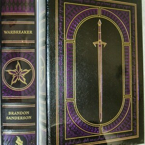 May include: A black leather-bound book with gold accents and a purple design. The title "Warbreaker" is printed on the spine. The book is by Brandon Sanderson.