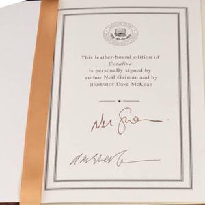 K&ouml;nnte beinhalten: Offenes Buch mit einem beigen Leseband. Die Seite zeigt den Text: &bdquo;Diese ledergebundene Ausgabe von Coraline ist vom Autor Neil Gaiman und vom Illustrator Dave McKean pers&ouml;nlich signiert.&ldquo; Zwei Unterschriften sind sichtbar.