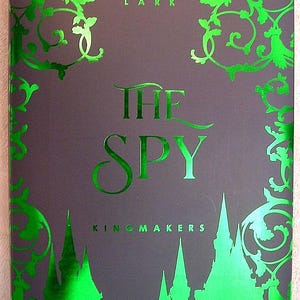 Może przedstawiać: Okładka książki "The Spy" autorstwa Sophie Lark. Ciemnoszare tło z jasnymi zielonymi akcentami. Tytuł i nazwisko autora są zielone, wraz z dekoracyjnymi wzorami i sylwetką zamku.