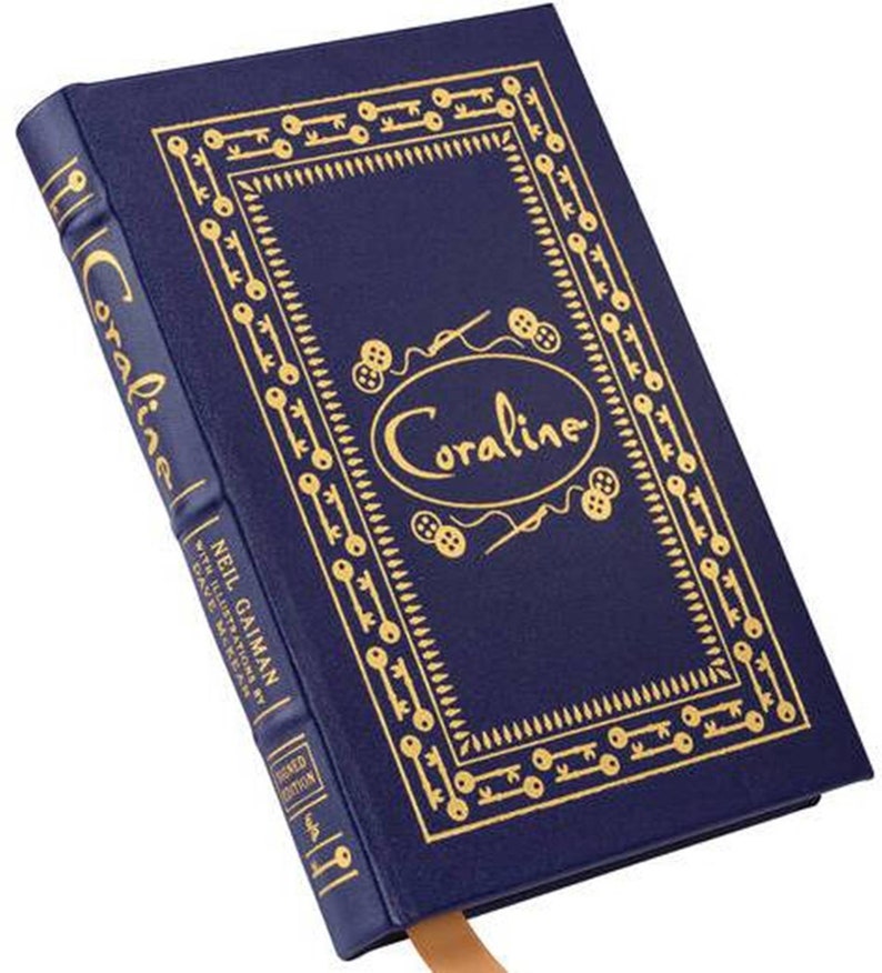 K&ouml;nnte beinhalten: Ein dunkelblaues Hardcover-Buch mit dem Titel "Coraline" und goldener Schrift. Der Einband zeigt einen dekorativen Rahmen mit Schl&uuml;ssel- und Knopffmotiven. Der Buchr&uuml;cken zeigt den Namen des Autors, Neil Gaiman. Ein Lesezeichen ist sichtbar.