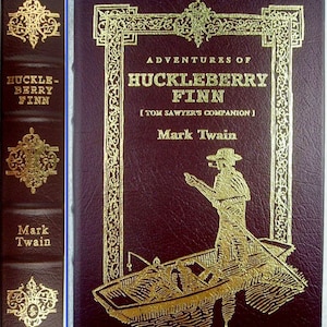 May include: A dark maroon hardback book titled Adventures of Huckleberry Finn by Mark Twain. The cover features gold foil lettering and an illustration of a person in a boat. The spine includes gold decorative filigree and the author's name.