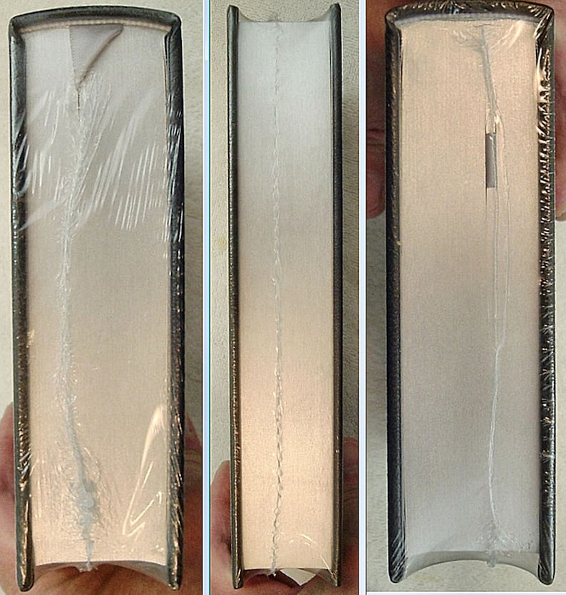 May include: Close-up of the spine of a hardcover book with a white cover and black binding. The book is wrapped in clear plastic.