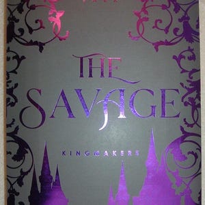 Può includere: Copertina di un libro con il titolo "The Savage" in grandi lettere metalliche viola su sfondo grigio. Il nome dell'autore, Sophie Lark, è in alto. Il fondo presenta "Kingmakers" e una silhouette di castello viola.