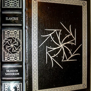 May include: A black leather-bound book with a silver embossed design on the cover. The title "ELANTRIS" is printed in silver on the spine, along with the author's name "BRANDON SANDERSON".