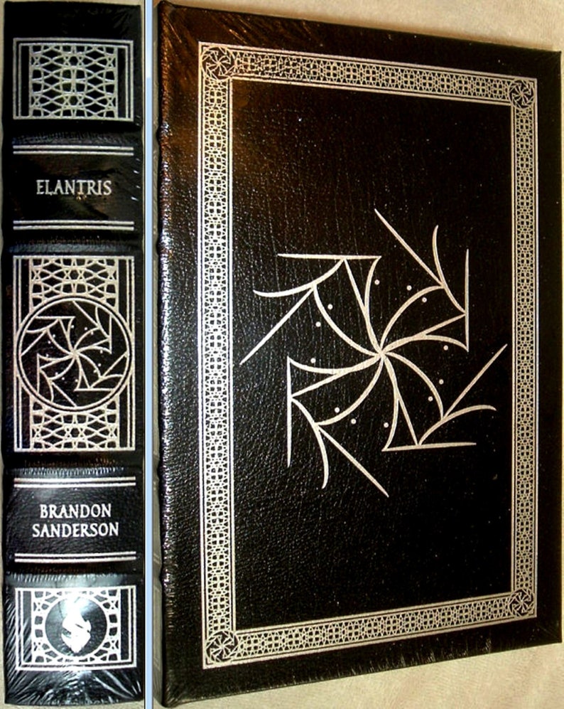 May include: A black leather-bound book with a silver embossed design on the cover. The title "ELANTRIS" is printed in silver on the spine, along with the author's name "BRANDON SANDERSON".