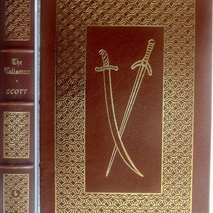 May include: A brown leather-bound book with gold accents. The title "The Talisman" and the author "SCOTT" are printed on the spine. The front cover features a gold embossed design of two crossed swords.