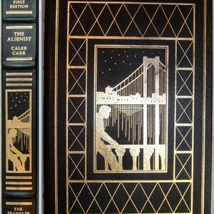 Op de afbeelding: Een hardcover boek met een zwarte kaft en gouden accenten. De kaft toont een gouden illustratie van een brug en een figuur. De rug leest "Signed First Edition" en "The Alienist" van Caleb Carr. Onderdeel van The Franklin Library.