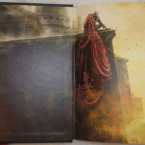 May include: A book cover with a dark brown background. The cover features a man in a black suit and a woman in a red dress standing on a ledge. The woman's dress has a floral pattern. The background is a cityscape with a building in the distance.