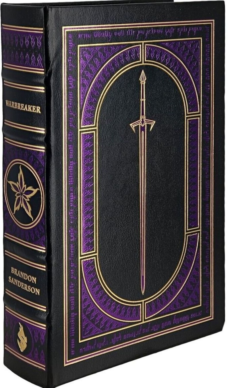 May include: A black leather-bound book with gold accents and a purple design. The title "Warbreaker" is printed on the spine. The front cover features a gold-framed oval with a sword inside.