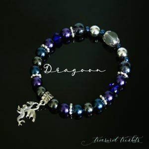 May include: A beaded bracelet with a silver dragon charm. The bracelet is made with dark blue, purple, and silver beads. The text "Dragoon" is visible in the center of the image.