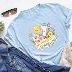 Peut inclure: T-shirt bleu clair avec l'inscription "Sunshine & Sonograms". Le motif comprend un soleil souriant, des fleurs et une sonde d'échographie. Des lunettes de soleil et une veste en jean sont en arrière-plan.