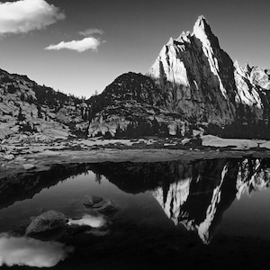 May include: Black and white landscape photograph of a mountain range reflected in a lake. The image features a sharp, jagged mountain peak, clouds, and a serene water surface, creating a dramatic and peaceful scene.